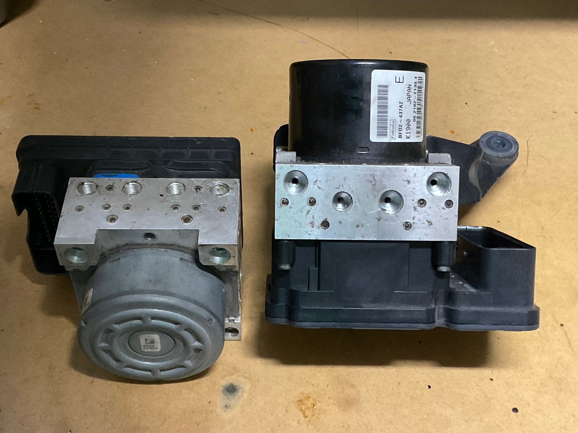 Two small metal mechanical parts with black housings on a cardboard surface, one showing a round front gear.