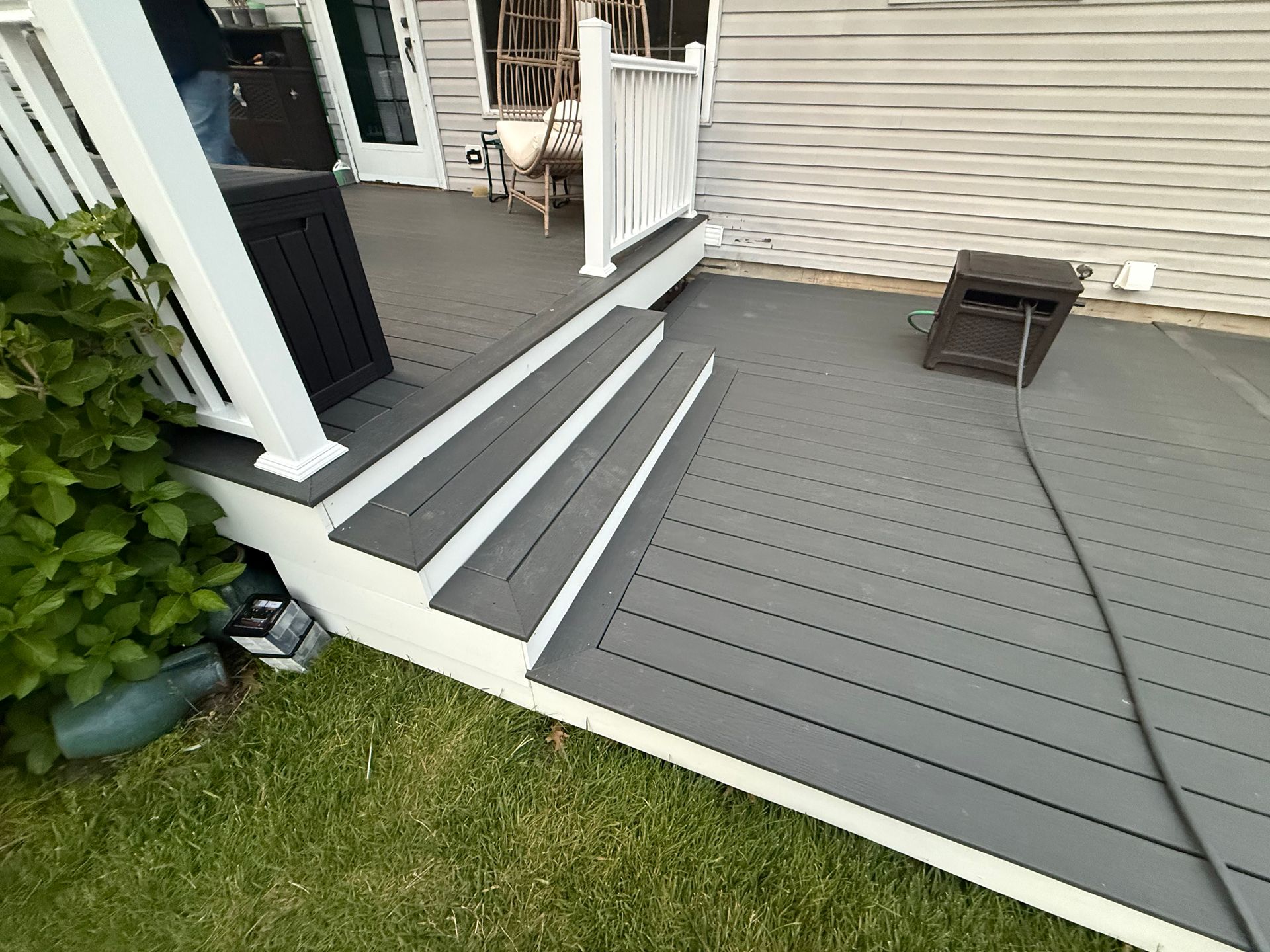Gray deck with steps leading to a porch beside a house, next to a grassy yard and a small stool.