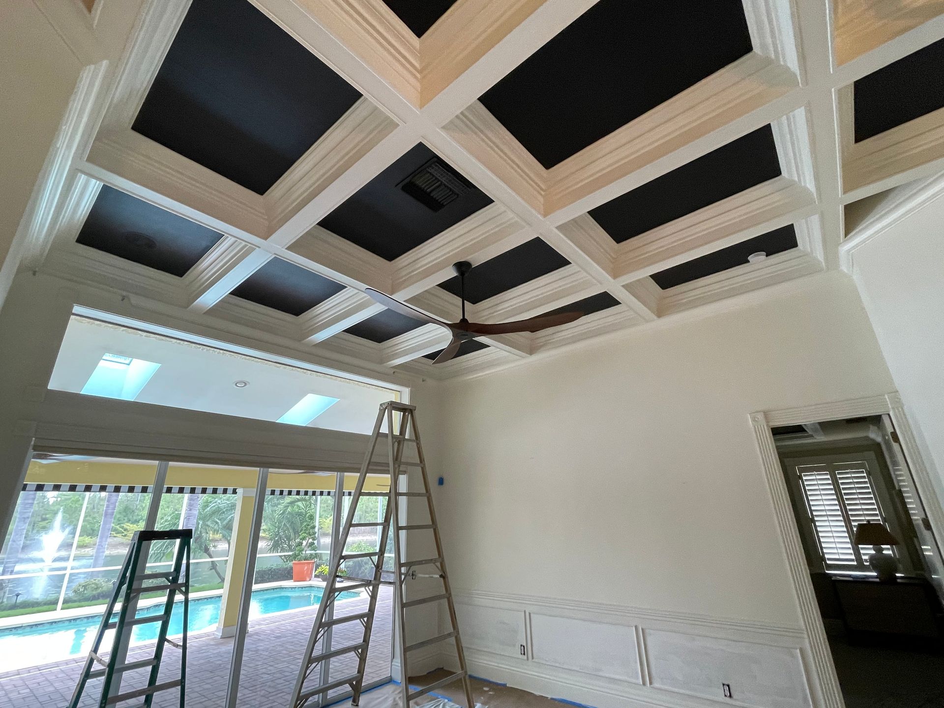 Interior room under construction with coffered ceiling, ladders, and large windows.