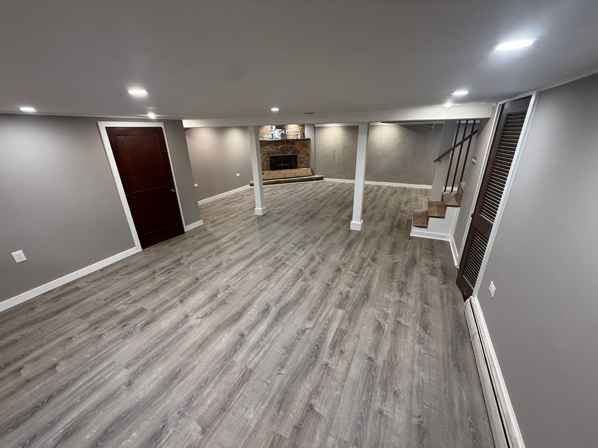 Empty finished basement with gray flooring, white columns, stairs, and recessed ceiling lights.