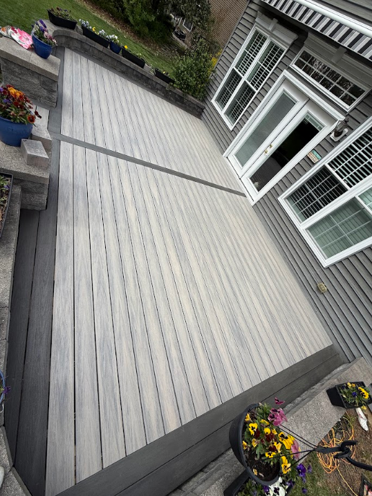 Large gray wooden deck beside a house with white windows and potted flowers.