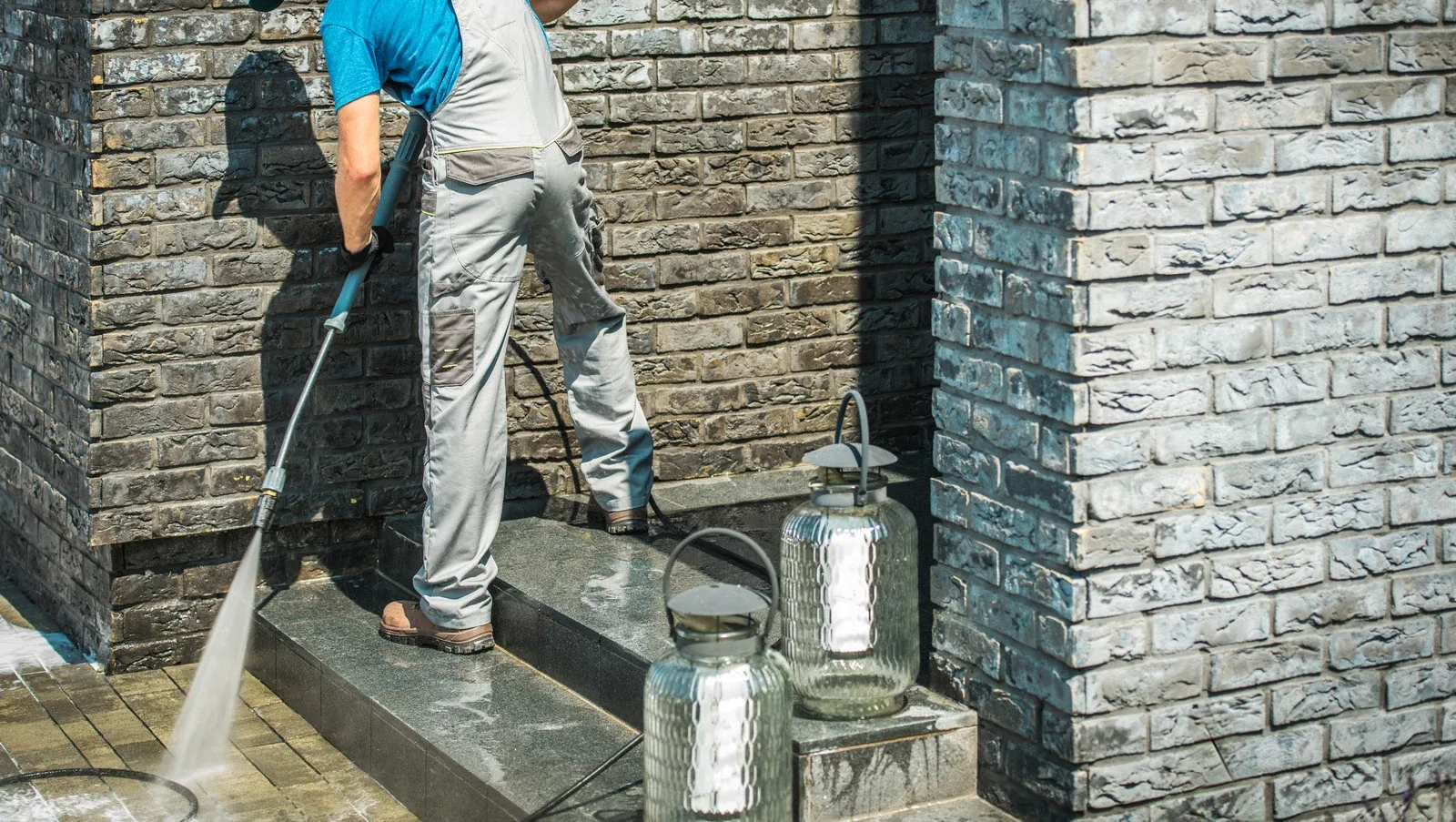 A person wearing light-colored work overalls uses a pressure washer to clean a brick wall and steps outdoors.