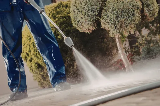 A person in blue waterproof gear uses a pressure washer to clean a paved surface outdoors near some shrubs.