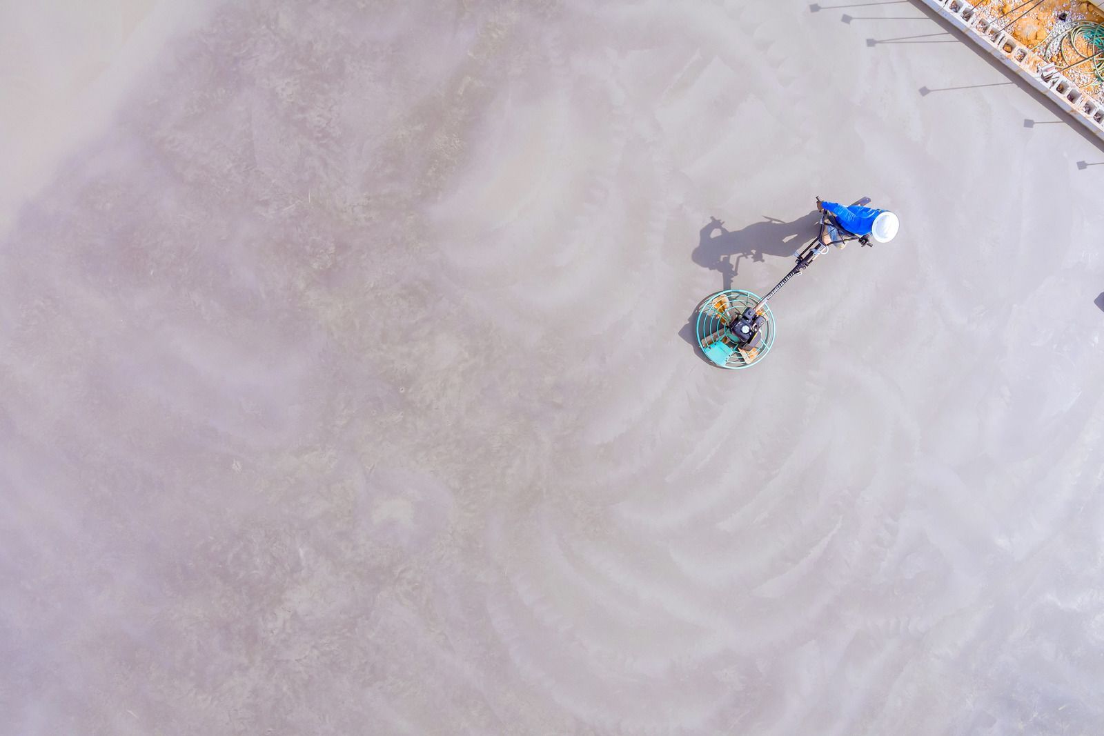 An aerial view of a worker using a power trowel to smooth a large, wet concrete surface.