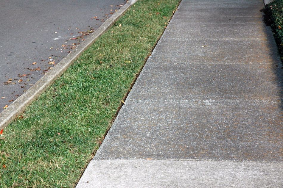 A gray concrete sidewalk runs parallel to a narrow strip of green grass and an asphalt road.