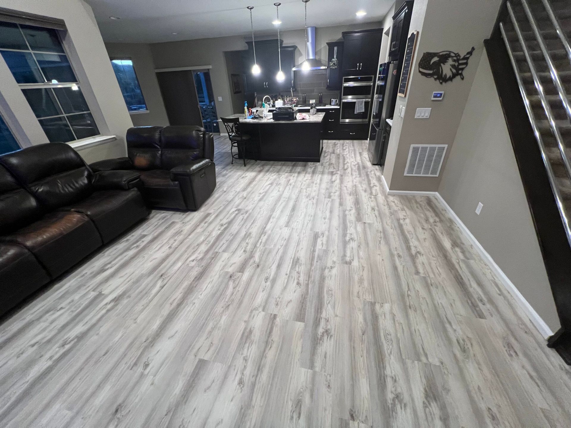 Modern open-plan living room with black leather sectional, gray wood floors, and kitchen island in the background