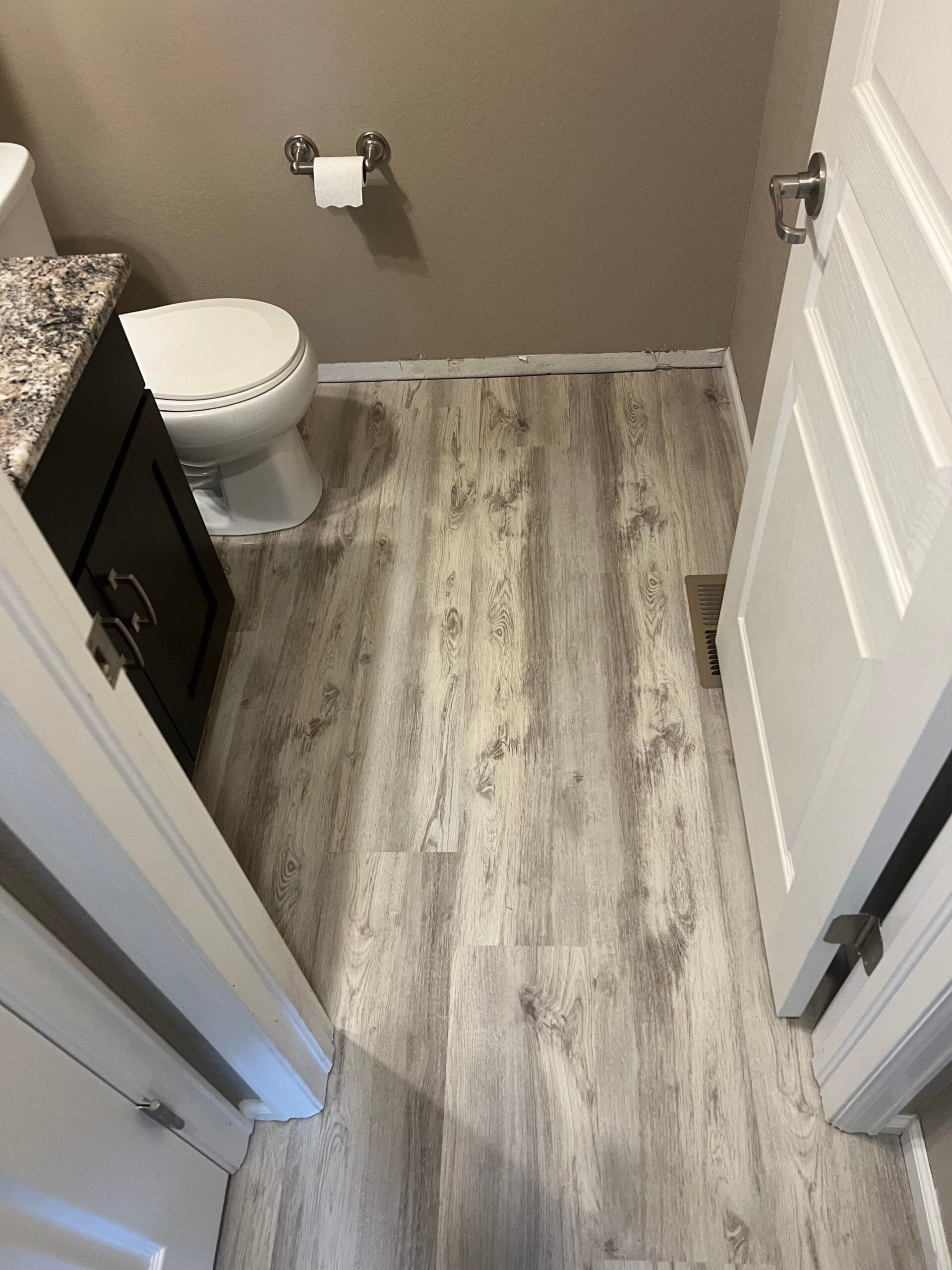 Small bathroom with wood-look flooring, white toilet, dark vanity, and partially open door.