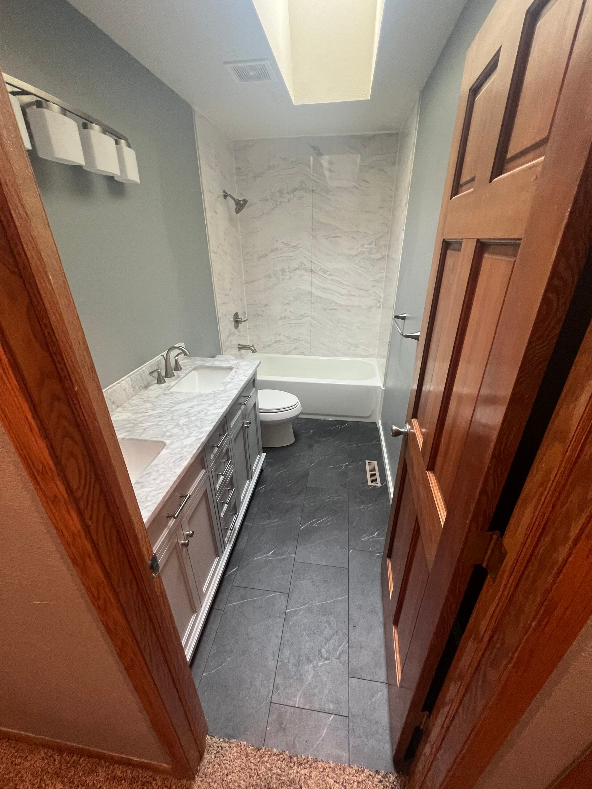 Narrow bathroom with white tub and vanity, gray tile floor, and wooden door open inward.
