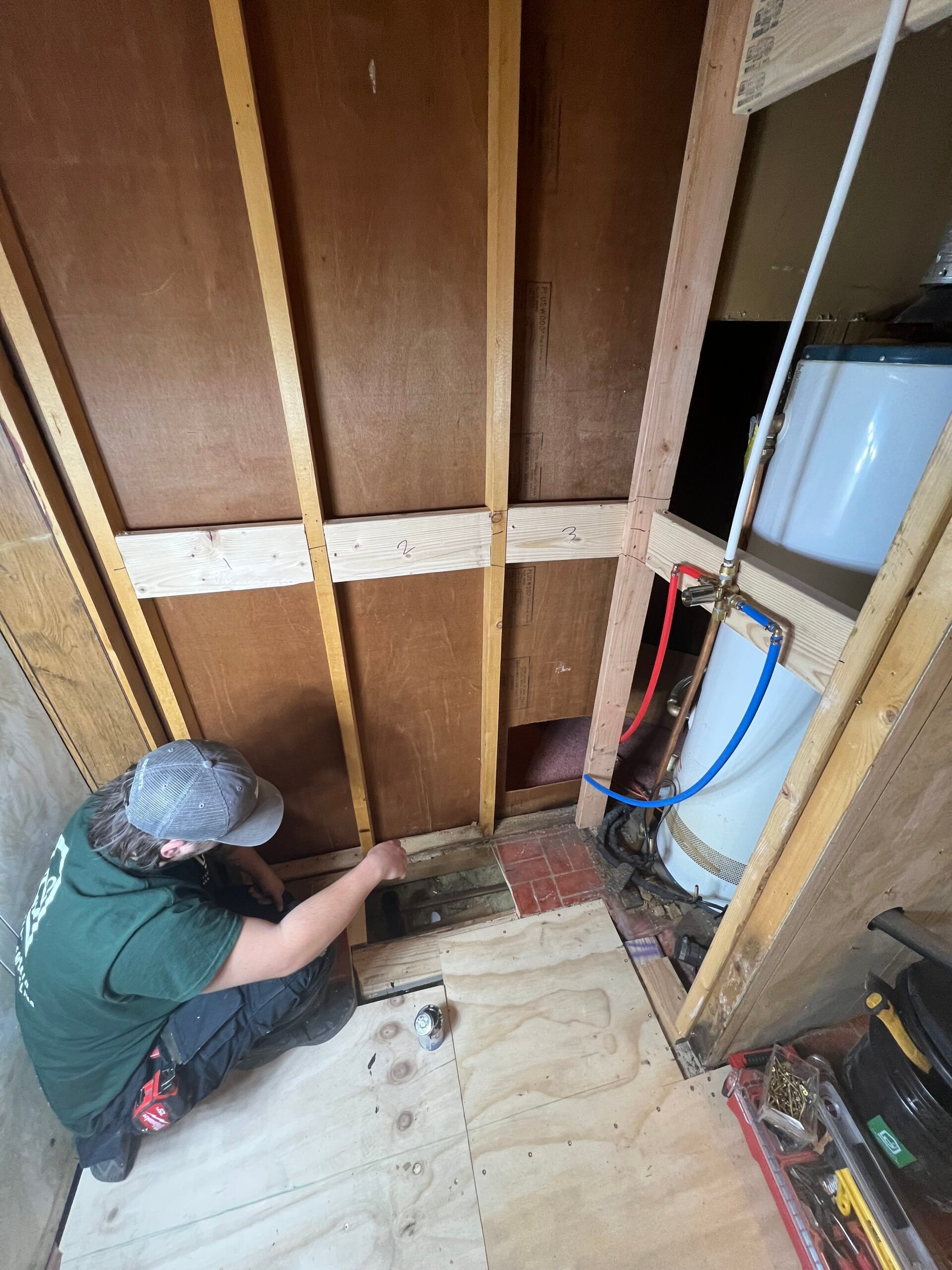 Person kneeling in an unfinished utility closet, working near plumbing and exposed wall studs.