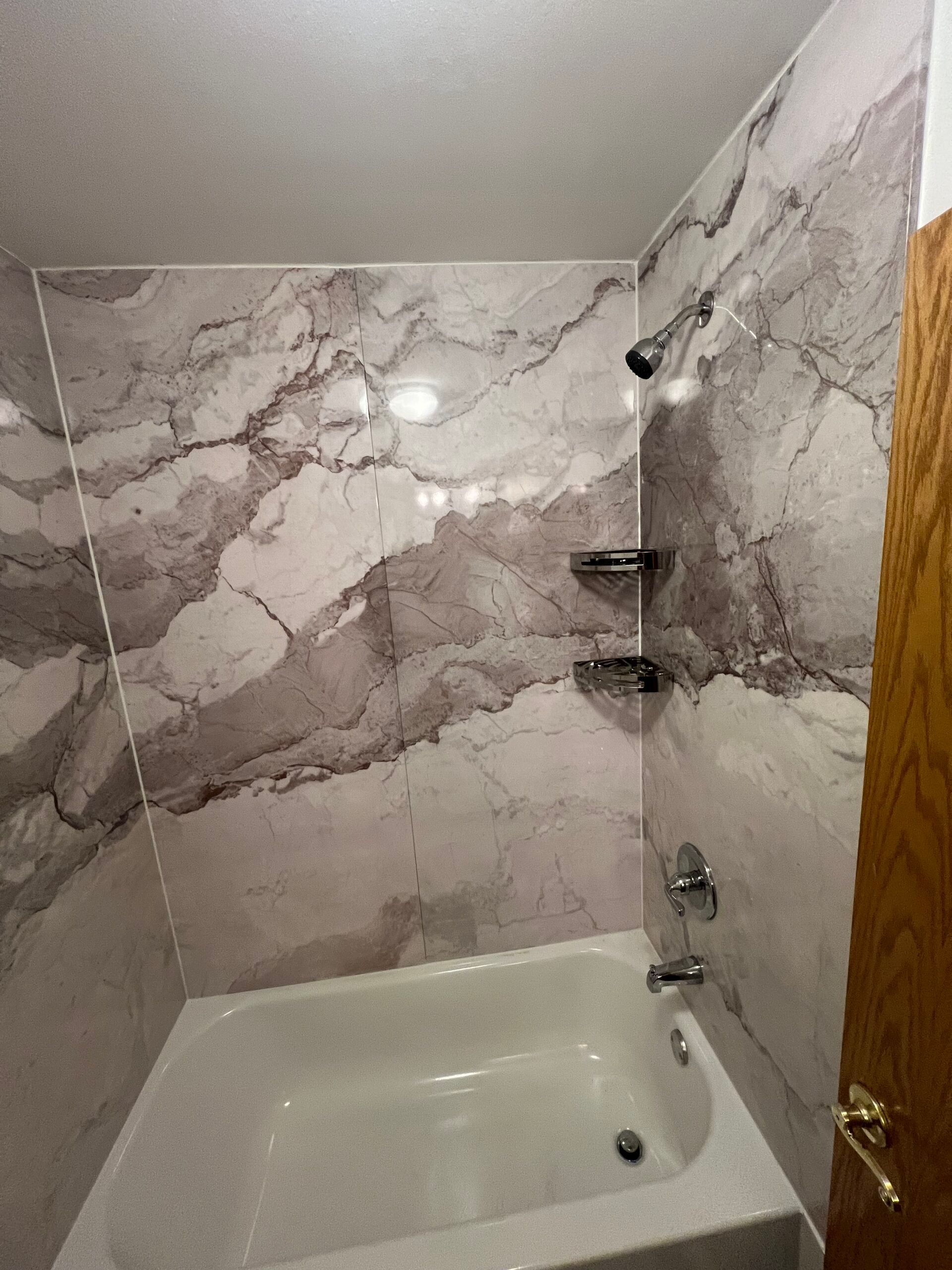 Small bathroom with marble-pattern shower walls, bathtub, and built-in corner shelves.
