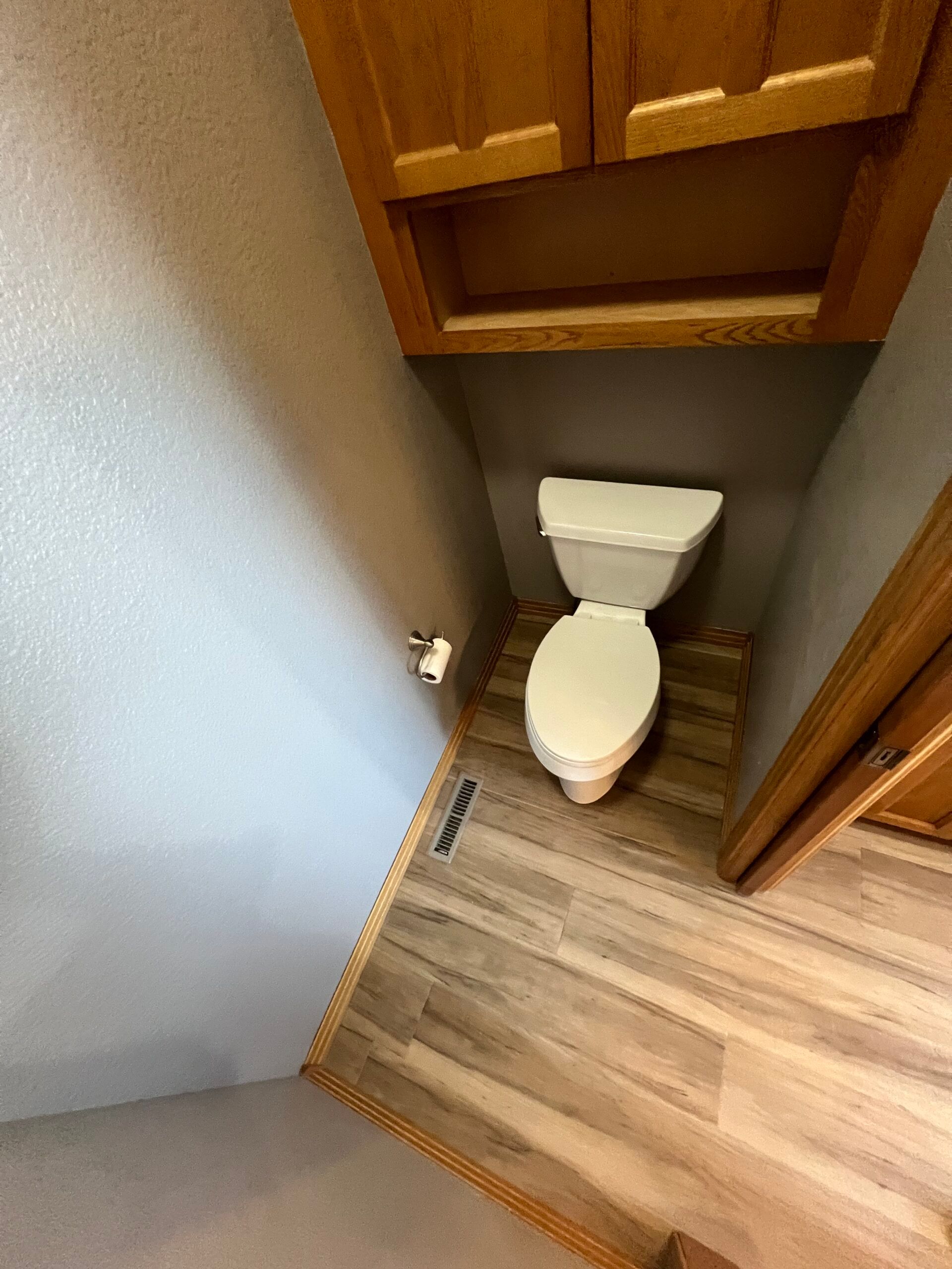 Small bathroom with a toilet tucked into a corner beside wood cabinets and laminate flooring.