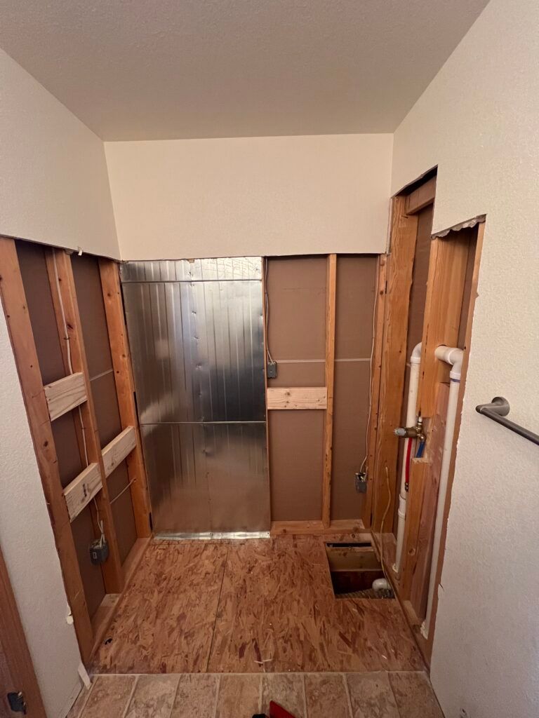 Partially gutted room with exposed wood framing, plywood floor, and metallic wall at the far end.