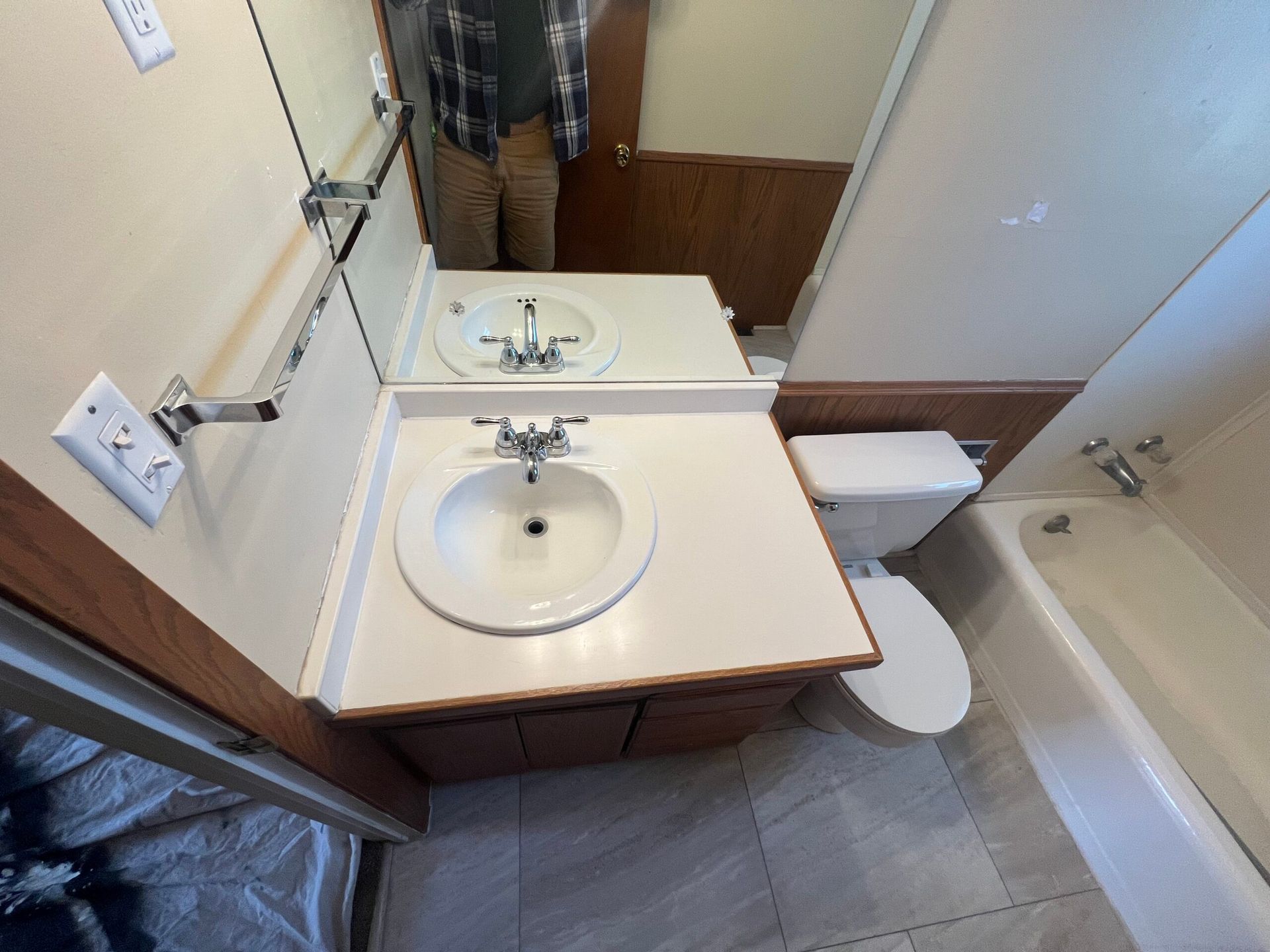 Small bathroom with sink vanity, mirror, toilet, and bathtub; person visible in mirror reflection.