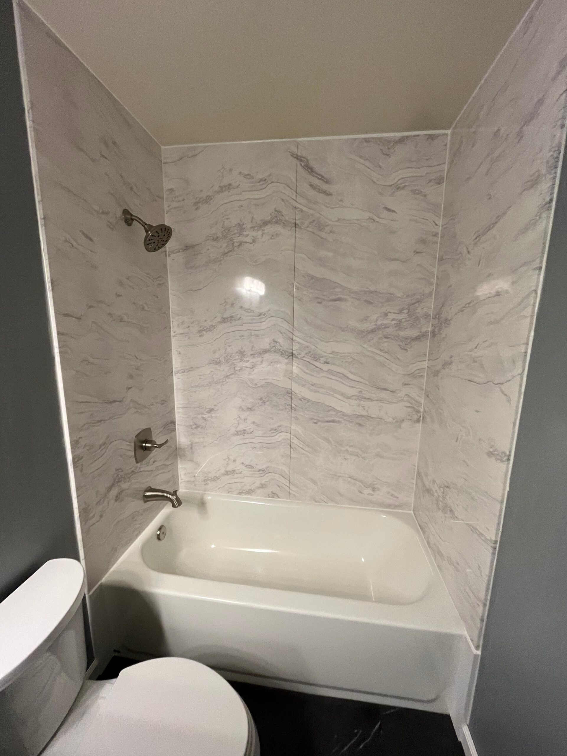 Small bathroom tub and shower with marble-look tile walls and a toilet in the foreground