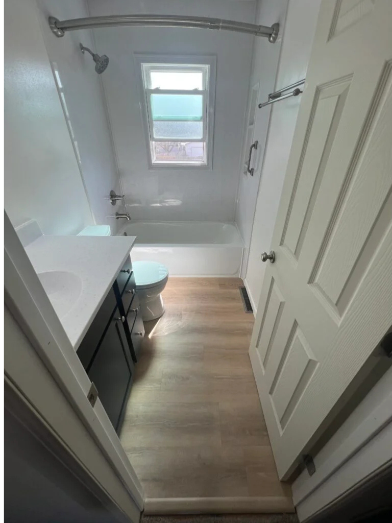 Small bathroom with white tub, toilet, vanity, and wood-look floor viewed from the doorway