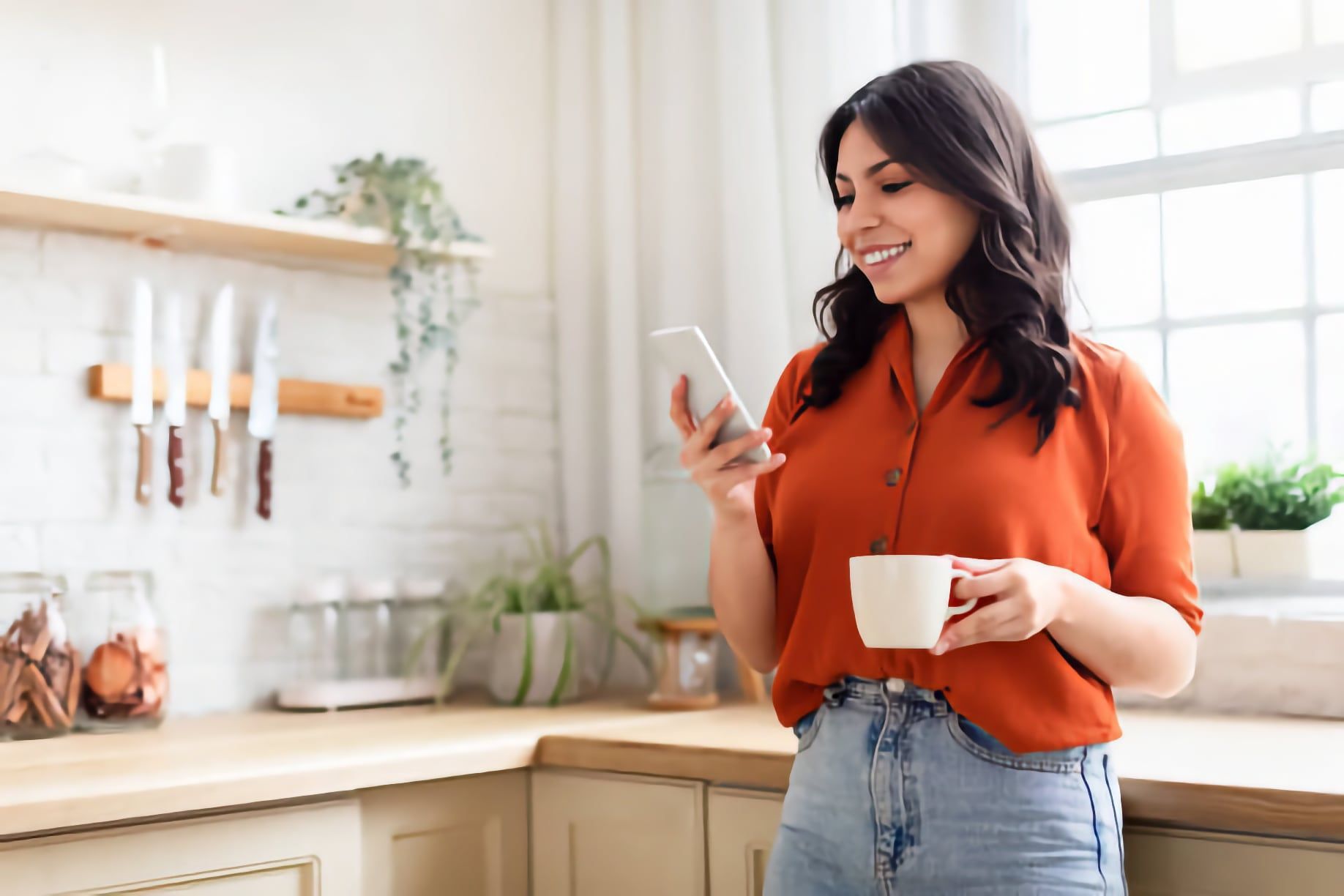 Woman in an orange blouse smiling at her phone while holding a mug in a bright kitchen