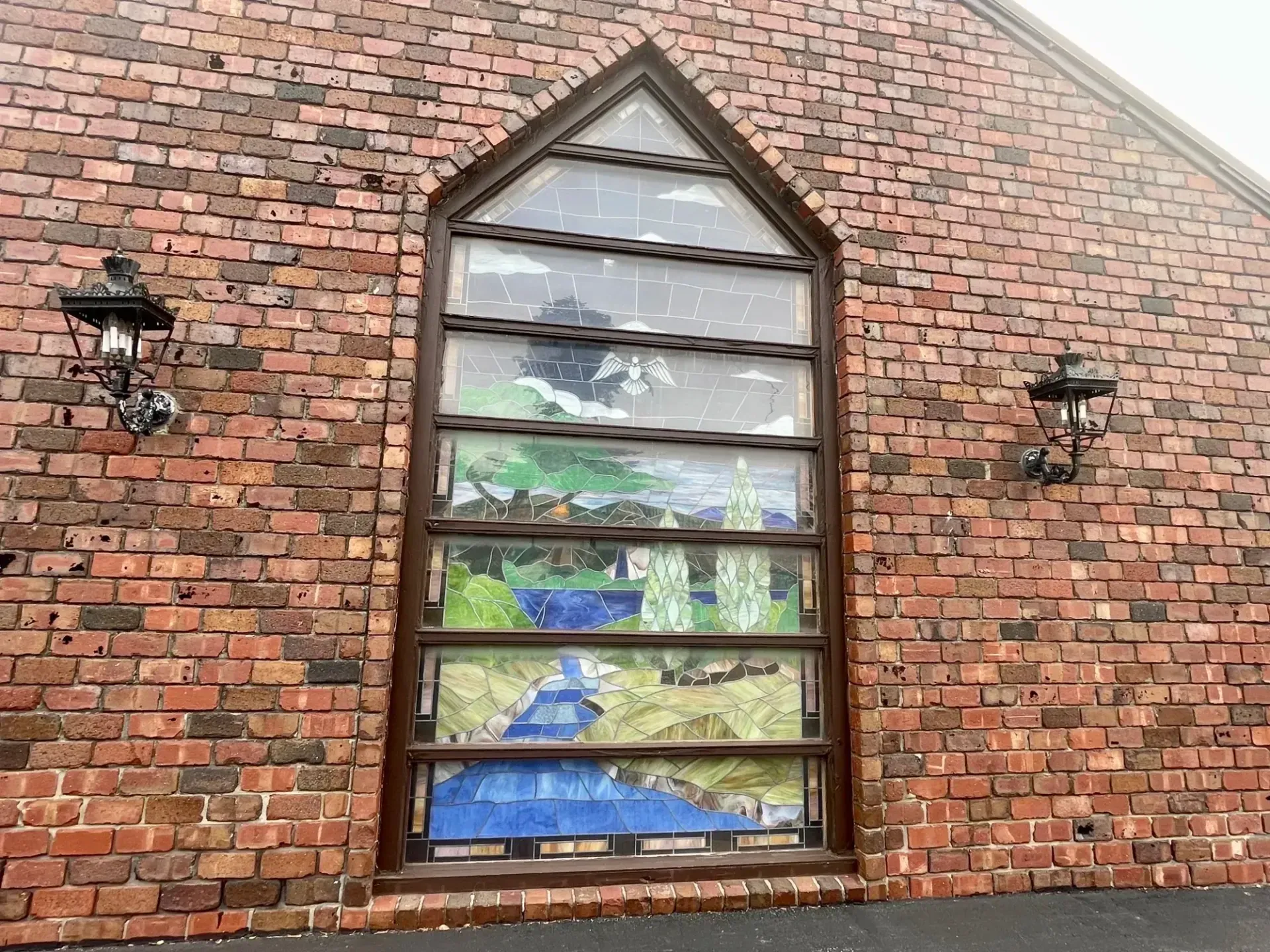 Brick wall with a tall arched stained-glass window and two black lanterns on either side.