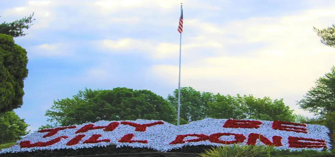 Flower bed spelling “WELCOME HOME” beneath an American flag on a landscaped hilltop.