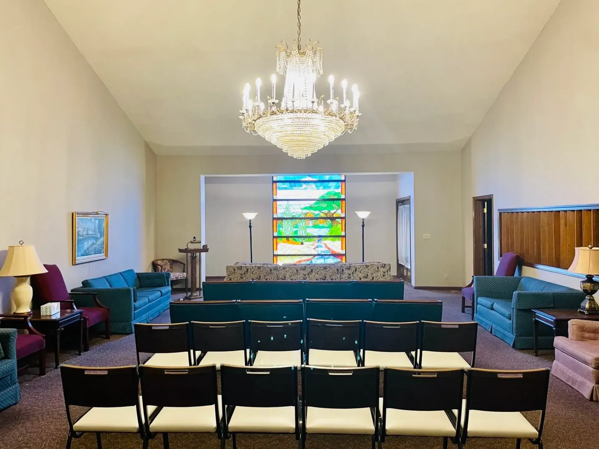 Empty chapel with rows of black chairs, a large chandelier, and a stained-glass window at the front.