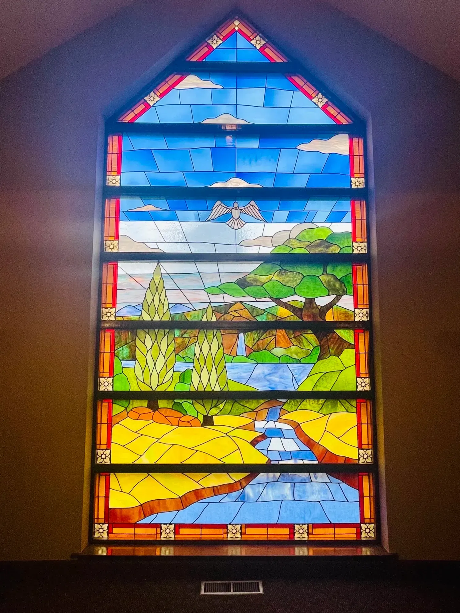 Colorful stained-glass church window showing a landscape with mountains, sky, water, and a sunlit field