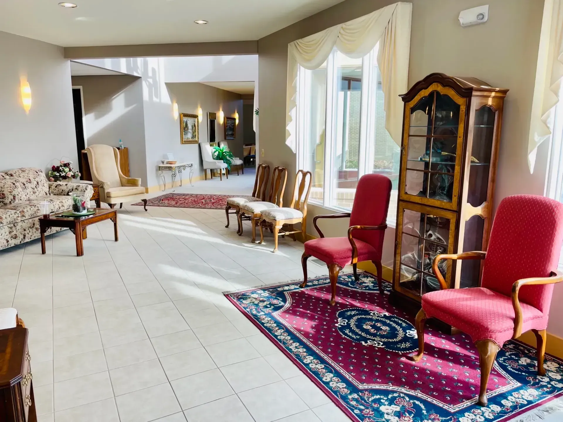 Bright living room with red chairs, patterned rug, wood cabinet, and sunlight through windows