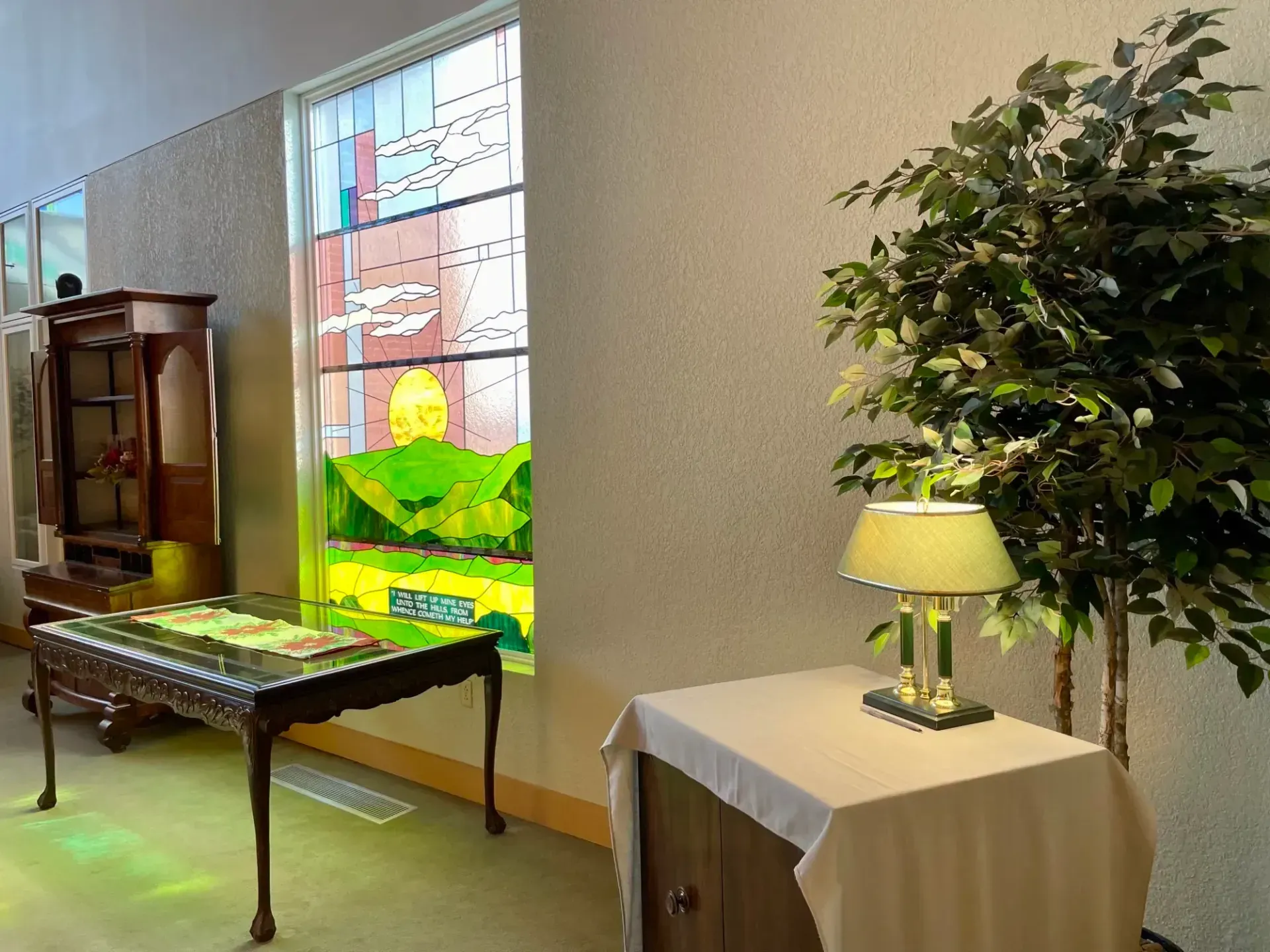 Church hallway with wooden table, stained-glass window, lamp on side table, and potted tree.