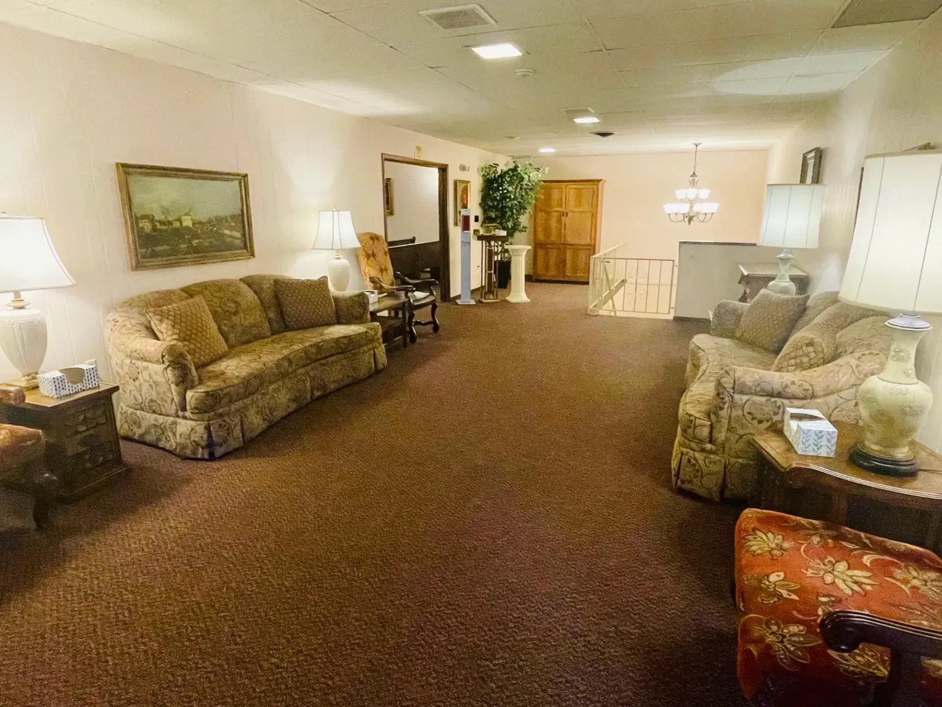 Spacious beige lobby with sofas, lamps, and a central carpeted walkway toward a wooden door.