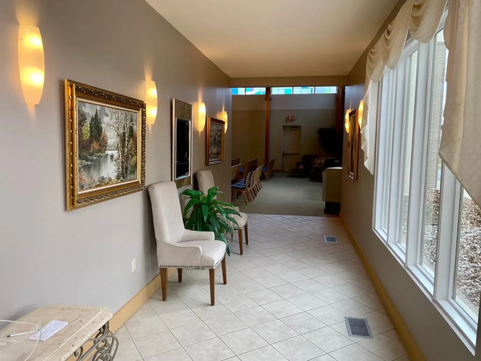 Long hotel hallway with white chairs, framed art, potted plants, and windows along the right side