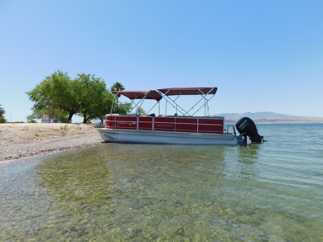 Red canopy boat moored in clear shallow water by a pebble shore and trees under a blue sky