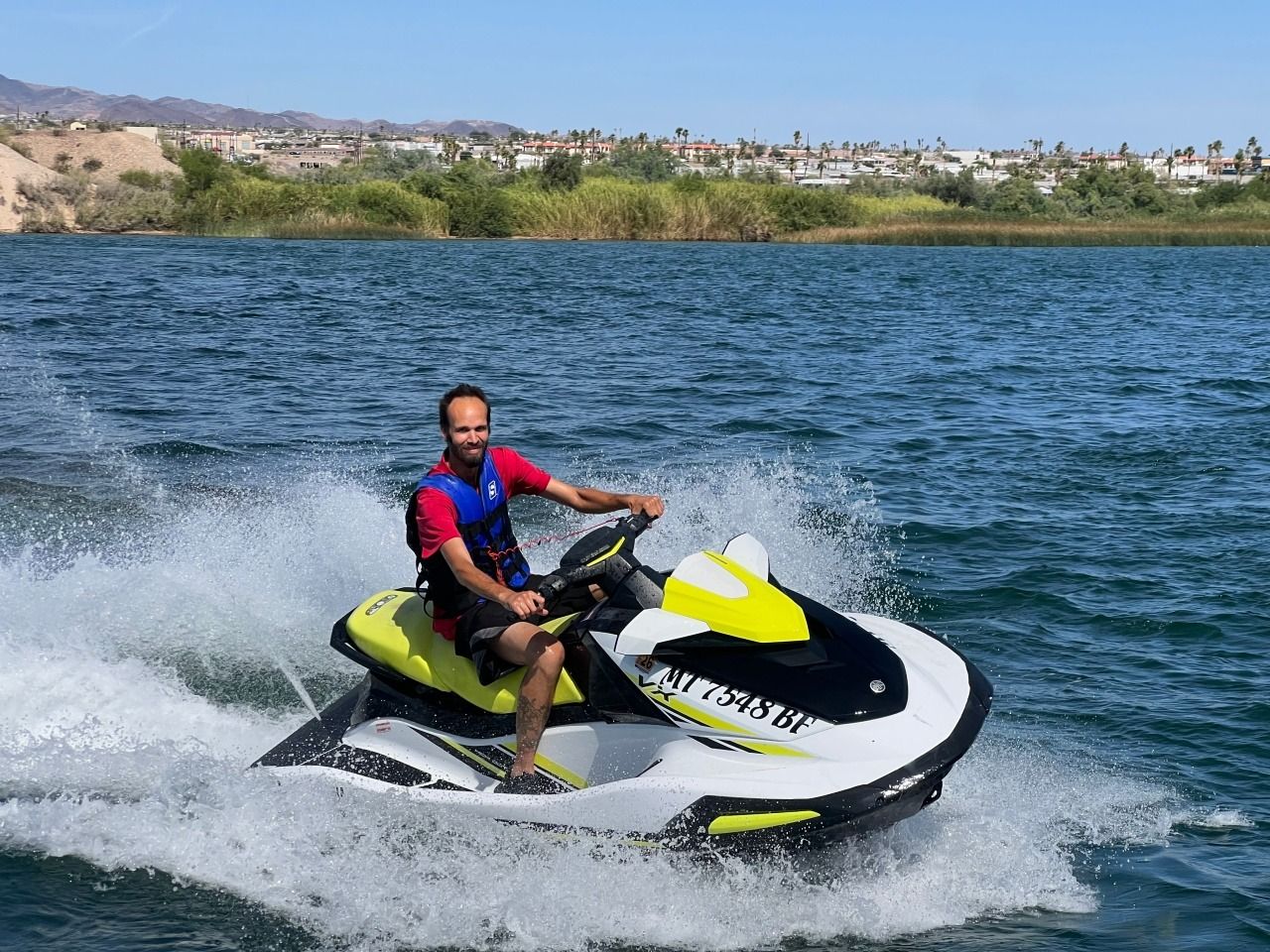 Person riding a yellow-and-black jet ski across a lake, splashing water with a shoreline in the background