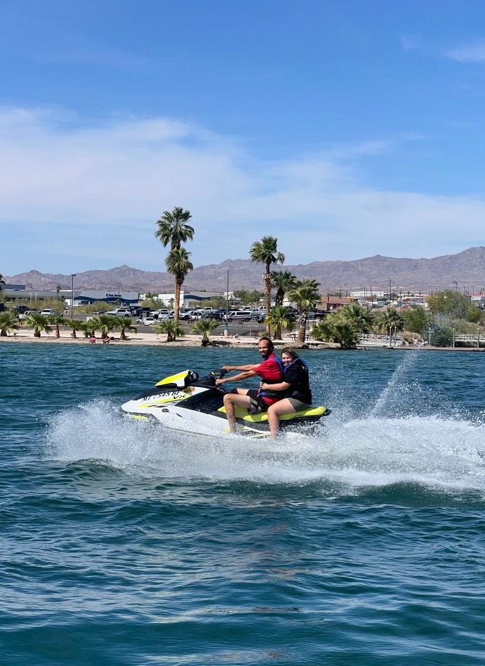 Person riding a jet ski across choppy blue water near a palm-lined shore and mountains.