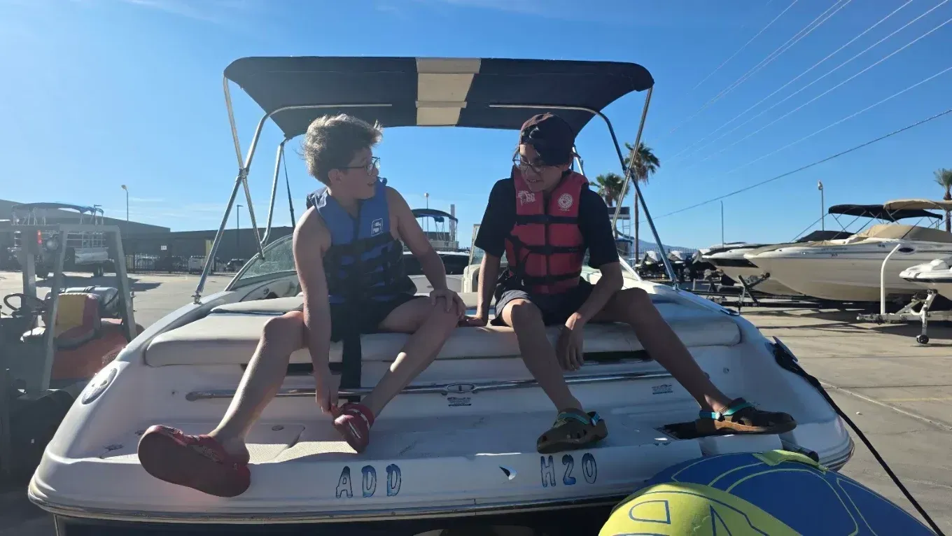 Two people sit on the back of a boat at a marina on a sunny day, wearing life jackets.
