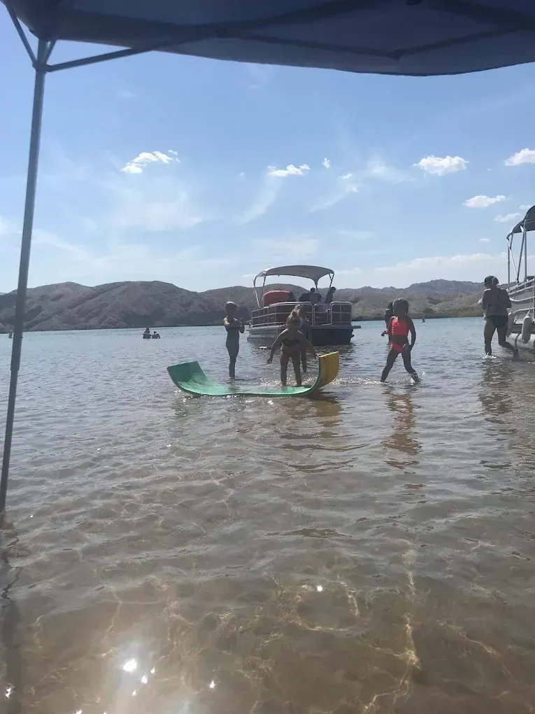 People wade by a small boat in clear shallow water near a rocky shoreline under a bright sky
