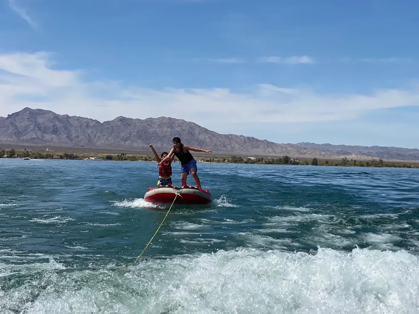Two people ride a red inflatable tube across choppy blue water with mountains in the background