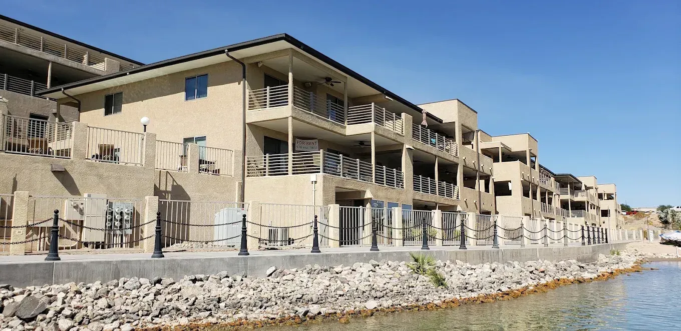 Waterfront row of beige apartments with balconies beside a rocky shoreline under a clear blue sky