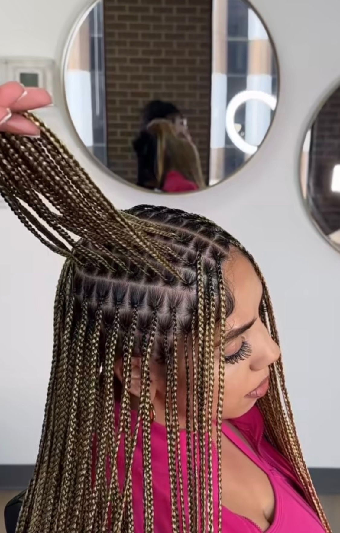 Woman getting blonde box braids in a salon, hand holding the braids, with a pink top.