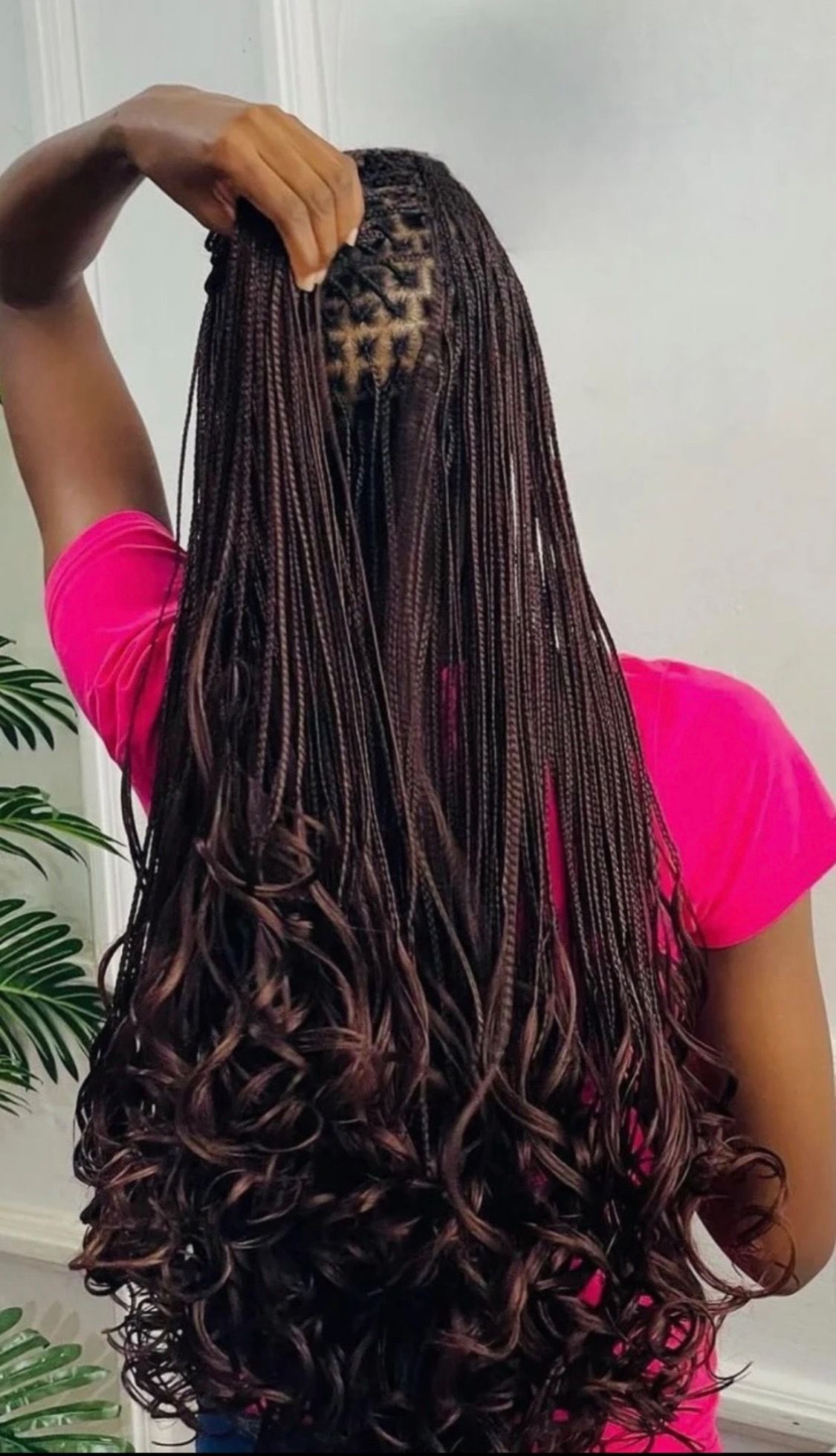 Woman with long, brown micro braids, some ends are curly, wearing a pink shirt, hand up, against a white wall.