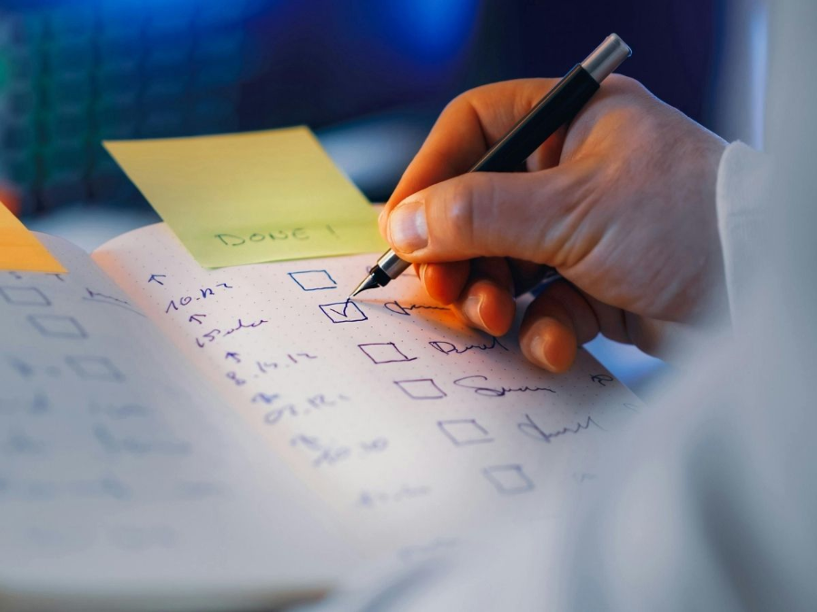 Hand checking boxes on a paper checklist with a pen beside a sticky note and notebook