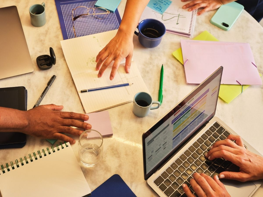 Team collaborating around a laptop with papers, notebooks, and coffee on a desk