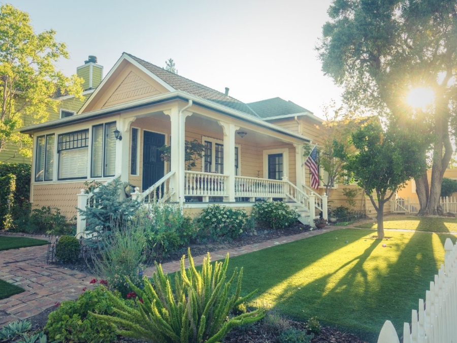 Sunlit cottage with a front porch, green lawn, and white picket fence in a leafy yard.