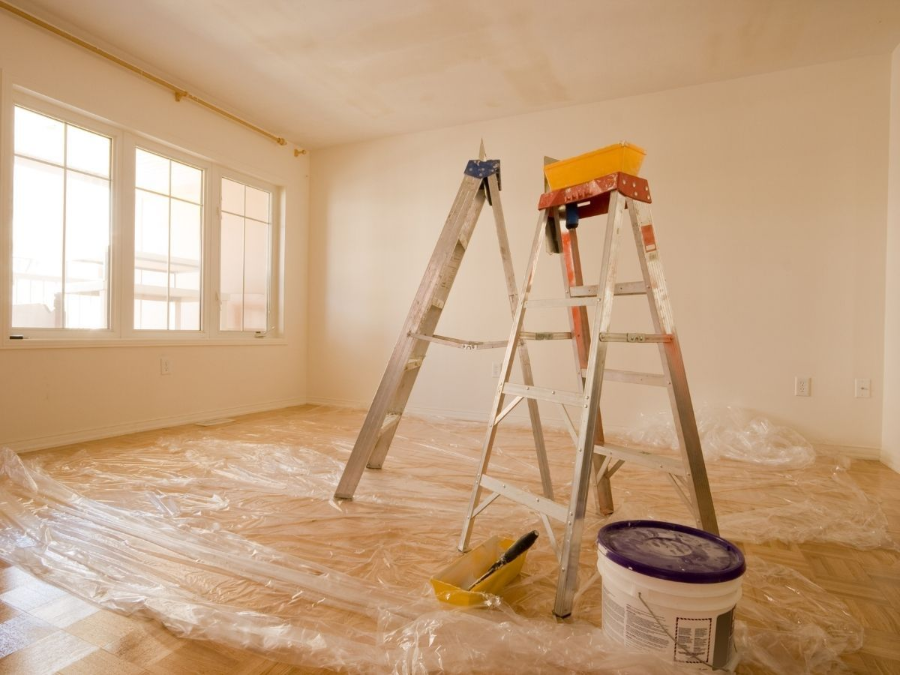Empty room being painted, with ladders, plastic-covered floor, and a window letting in light.