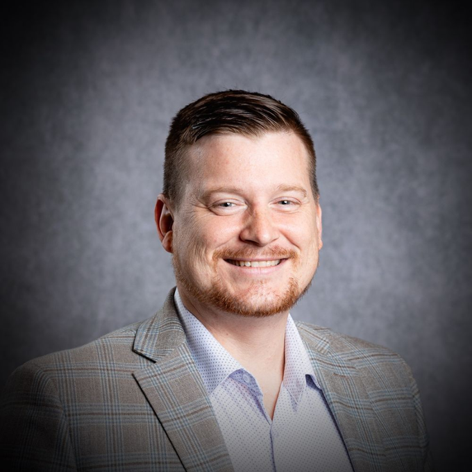 Smiling man in a gray plaid blazer against a dark studio background