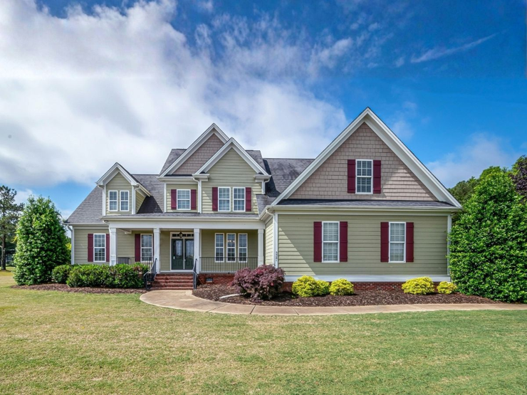 Large beige suburban house with red shutters, front porch, and a manicured lawn under a blue sky