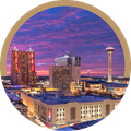 City skyline at sunset with colorful clouds, lit buildings, and a tall observation tower.