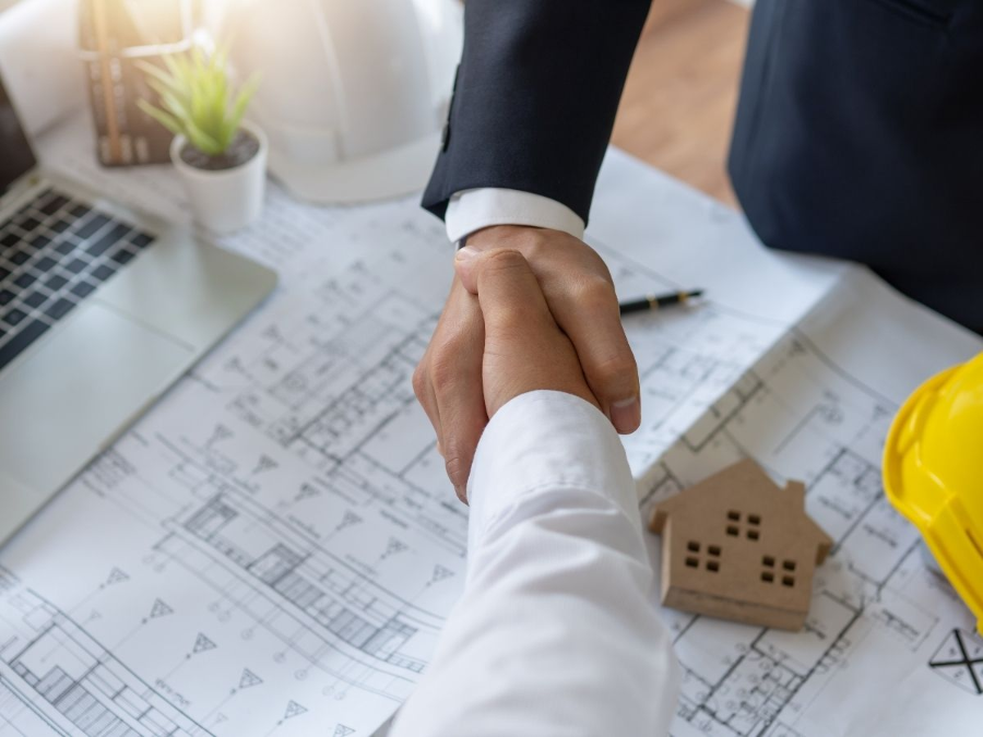 Handshake over architectural blueprints beside a small house model and yellow hard hat.