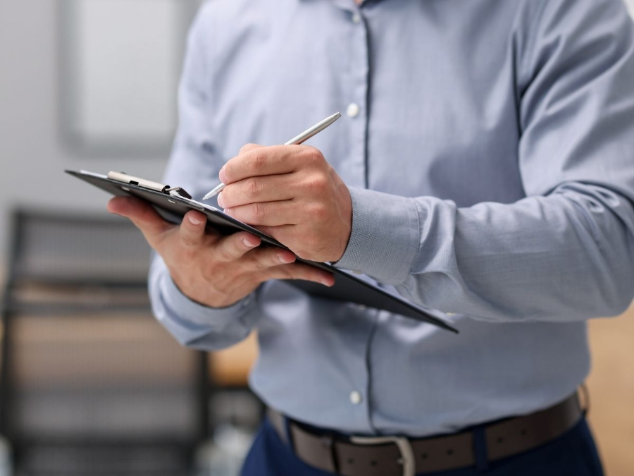Person in a blue shirt writing on a clipboard with a pen in an office setting