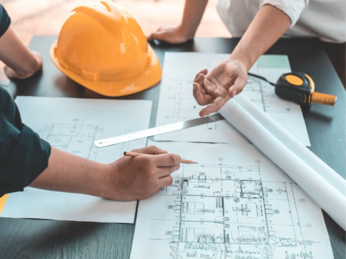 Hands reviewing blueprints on a desk with an orange hard hat and measuring tools