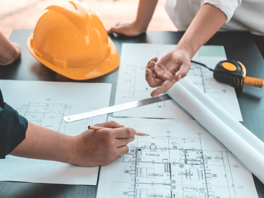 Hands reviewing architectural blueprints with a yellow hard hat and measuring tape on a desk.