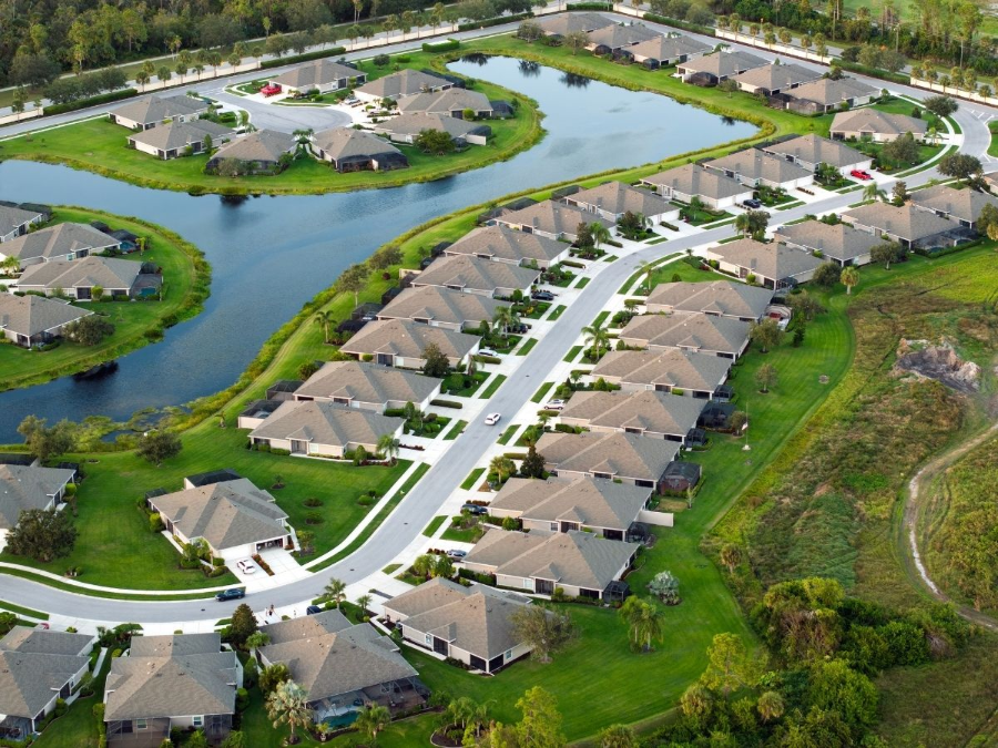 Aerial view of a suburban neighborhood with tan-roofed houses, winding roads, and a lake/pond.