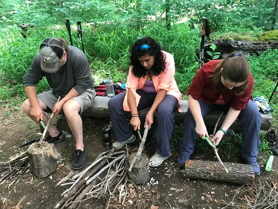 Three people are sitting on a bench in the woods cutting wood.